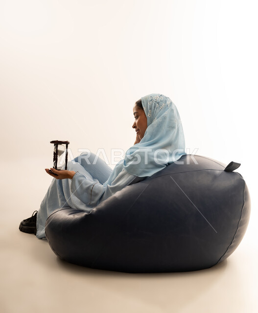 الجلوس بتعابير الملل والانتظار والسؤال, إدارة الأعمال وتنظيم الجدول الزمني, القيام بمراقبة توقيت الرمل, إدراك قيمة اللحظات الثمينة, بورتريه لشابة عربية خليجية عمانيه ترتدي العباءة الملونة والحجاب تمسك بيدها الساعة الرملية الزجاجية (البنكام), خلفية بيضاء