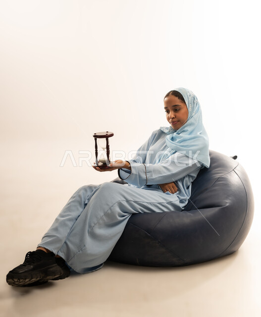الجلوس بتعابير الملل والانتظار والسؤال, إدارة الأعمال وتنظيم الجدول الزمني, القيام بمراقبة توقيت الرمل, إدراك قيمة اللحظات الثمينة, بورتريه لشابة عربية خليجية عمانيه ترتدي العباءة الملونة والحجاب تمسك بيدها الساعة الرملية الزجاجية (البنكام), خلفية بيضاء