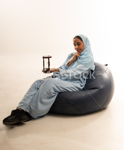 الجلوس بتعابير الملل والانتظار والسؤال, إدارة الأعمال وتنظيم الجدول الزمني, القيام بمراقبة توقيت الرمل, إدراك قيمة اللحظات الثمينة, بورتريه لشابة عربية خليجية عمانيه ترتدي العباءة الملونة والحجاب تمسك بيدها الساعة الرملية الزجاجية (البنكام), خلفية بيضاء