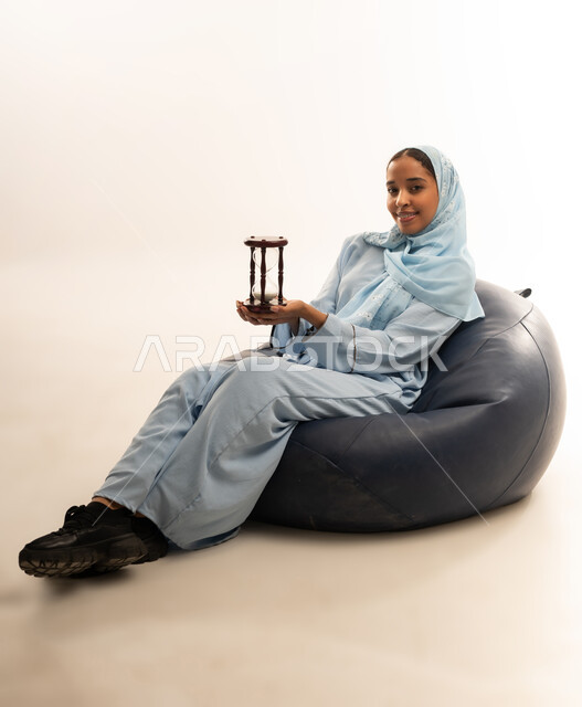الجلوس بتعابير الملل والانتظار والسؤال, إدارة الأعمال وتنظيم الجدول الزمني, القيام بمراقبة توقيت الرمل, إدراك قيمة اللحظات الثمينة, بورتريه لشابة عربية خليجية عمانيه ترتدي العباءة الملونة والحجاب تمسك بيدها الساعة الرملية الزجاجية (البنكام), خلفية بيضاء