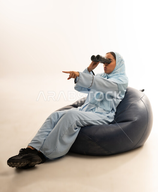 A vision of the future among young people, a love of exploration and adventure, gestures and facial expressions indicating surprise and astonishment, a portrait of an Arab Gulf Omani woman sitting on a comfortable chair looking through a viewfinder and pointing at something with her hand, white background