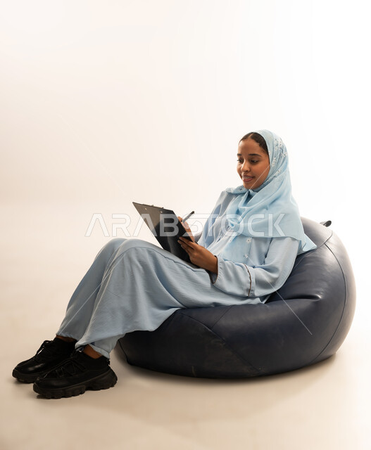 كتابة وتسجيل الملاحظات, التمعن والتركيز, إيماءات وجه ويدين تدل على التفكير, بورتريه  لسيده عربيه خليجيه عمانيه ترتدي العباءه الملونه تجلس على كرسي مريح تمسك بيدها لوح وقلم, خلفيه بيضاء