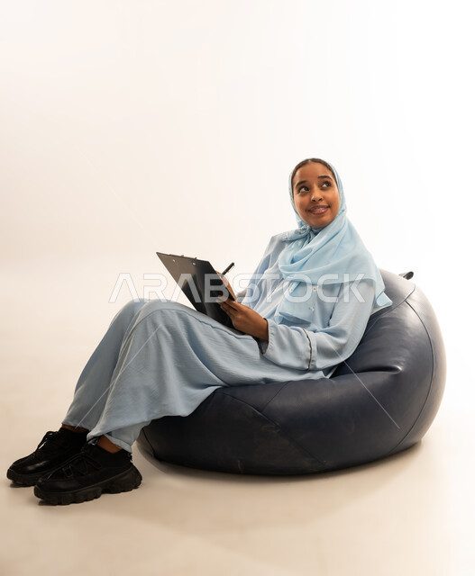 كتابة وتسجيل الملاحظات, التمعن والتركيز, إيماءات وجه ويدين تدل على التفكير, بورتريه  لسيده عربيه خليجيه عمانيه ترتدي العباءه الملونه تجلس على كرسي مريح تمسك بيدها لوح وقلم, خلفيه بيضاء