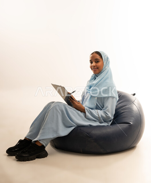 كتابة وتسجيل الملاحظات, التمعن والتركيز, إيماءات وجه ويدين تدل على التفكير, بورتريه  لسيده عربيه خليجيه عمانيه ترتدي العباءه الملونه تجلس على كرسي مريح تمسك بيدها لوح وقلم, خلفيه بيضاء