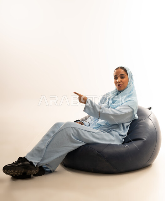 الجلوس على كرسي مريح والاشاره الى مكان ما، بورتريه لسيدة عربية خليجية عمانيه ترتدي العباءة الملونه والحجاب تشير باصبع السبابه باتجاه ما بايماءات السعاده والبهجه، حركات تدل على هذا المكان (هذا الموقع)، خلفية بيضاء