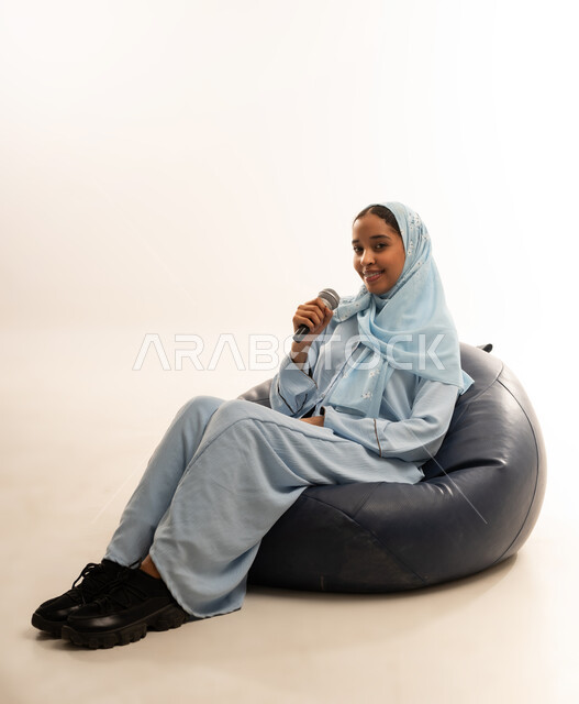 Using advanced technology and modern technical devices, a portrait of an Arab Gulf Omani girl wearing a colorful abaya sitting on a comfortable chair holding a microphone in her hand, gestures of happiness and pleasure in performing singing clips, white background