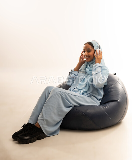 Using advanced technology and modern technical devices, portrait of an Arab Gulf Omani girl wearing a colorful abaya sitting on a comfortable chair with headphones on her head, gestures of happiness and pleasure listening to music, white background