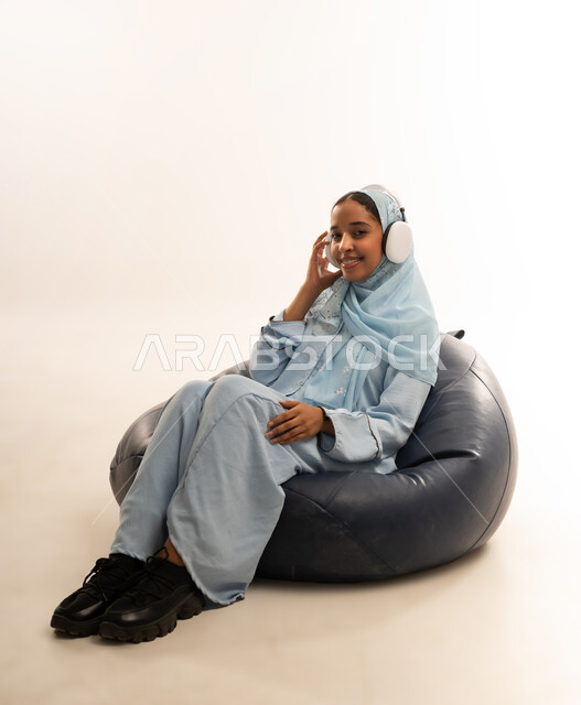 Using advanced technology and modern technical devices, portrait of an Arab Gulf Omani girl wearing a colorful abaya sitting on a comfortable chair with headphones on her head, gestures of happiness and pleasure listening to music, white background