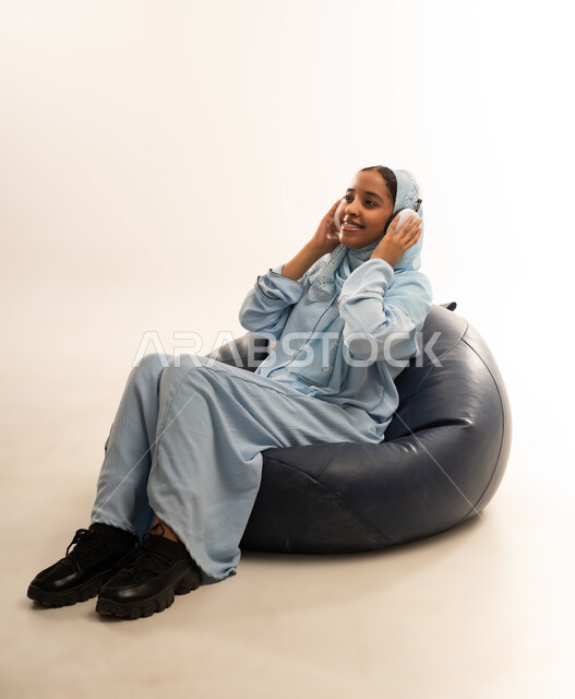 استخدام التكنولوجيا و الأجهزة التقنية الحديثة المتطورة, بورتريه لفتاة عربية خليجية عمانيه  ترتدي العباءة الملونه تجلس على كرسي مريح تضع سماعة الهيدفون حول رأسها, ايماءات السعادة والسرور بالاستماع الى الموسيقى, خلفية بيضاء