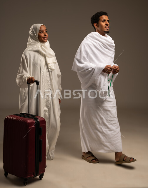 التسبيح والتهليل وذكر الله، التقرب الى الله بالعبادة والطاعة, الاستعداد لأداء مناسك الحج والعمرة، بورتريه بالطول الكامل لرجل وامرأه عربيان عمانيان خليجيان يرتديان ملابس الإحرام يمسك الرجل بيده السبحة ذات الحبات الخضراء وتمسك زوجته بيدها شنطه السفر، خلفية بيج