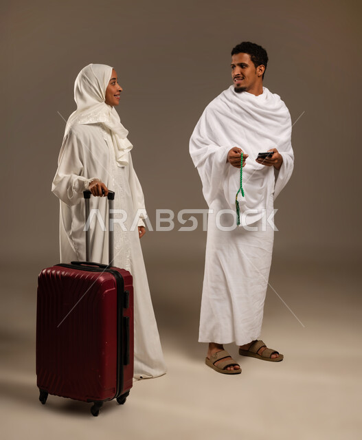 التسبيح والتهليل وذكر الله، التقرب الى الله بالعبادة والطاعة, الاستعداد لأداء مناسك الحج والعمرة، بورتريه بالطول الكامل لرجل وامرأه عربيان عمانيان خليجيان يرتديان ملابس الإحرام يمسك الرجل بيده السبحة ذات الحبات الخضراء وتمسك زوجته بيدها شنطه السفر، خلفية بيج