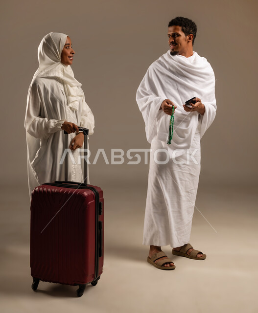 التسبيح والتهليل وذكر الله، التقرب الى الله بالعبادة والطاعة, الاستعداد لأداء مناسك الحج والعمرة، بورتريه بالطول الكامل لرجل وامرأه عربيان عمانيان خليجيان يرتديان ملابس الإحرام يمسك الرجل بيده السبحة ذات الحبات الخضراء وتمسك زوجته بيدها شنطه السفر، خلفية بيج