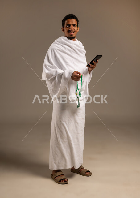 التسبيح والتهليل وذكر الله، التقرب الى الله بالعبادة والطاعة, أداء مناسك الحج والعمرة، بورتريه بالطول الكامل لرجل عربي عماني خليجي يرتدي ملابس الإحرام يمسك السبحة ذات الحبات الخضراء بيده وبيده الاخرى يقوم بتصفح هاتفهه المحمول، خلفية بيج