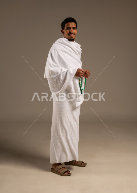 التسبيح والتهليل وذكر الله، التقرب الى الله بالعبادة والطاعة, أداء مناسك الحج والعمرة، بورتريه بالطول الكامل لرجل عربي عماني خليجي يرتدي ملابس الإحرام يمسك السبحة ذات الحبات الخضراء بيده ويسبح عبرها، خلفية بيج