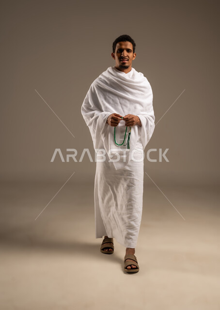 التسبيح والتهليل وذكر الله، التقرب الى الله بالعبادة والطاعة, أداء مناسك الحج والعمرة، بورتريه بالطول الكامل لرجل عربي عماني خليجي يرتدي ملابس الإحرام يمسك السبحة ذات الحبات الخضراء بيده ويسبح عبرها، خلفية بيج