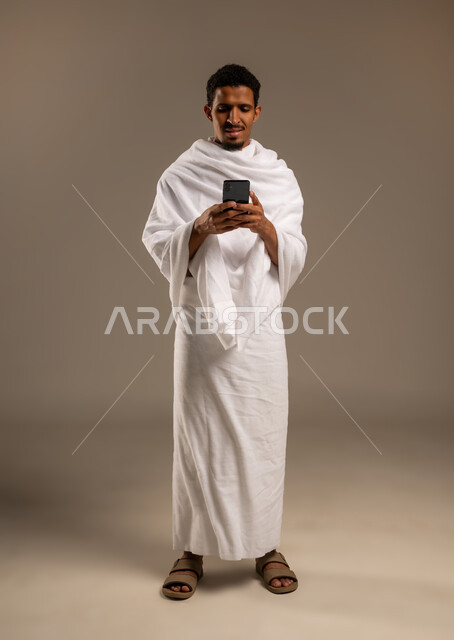 عرض تطبيقات وبرامج دليل الحاج والمعتمر، بورتريه لشاب خليجي عربي عماني يرتدي ثياب الإحرام  يمسك الهاتف المحمول، استخدام الأجهزة التقنية الحديثه في التواصل، صوره الجسم بالطول الكامل، خلفية بيج