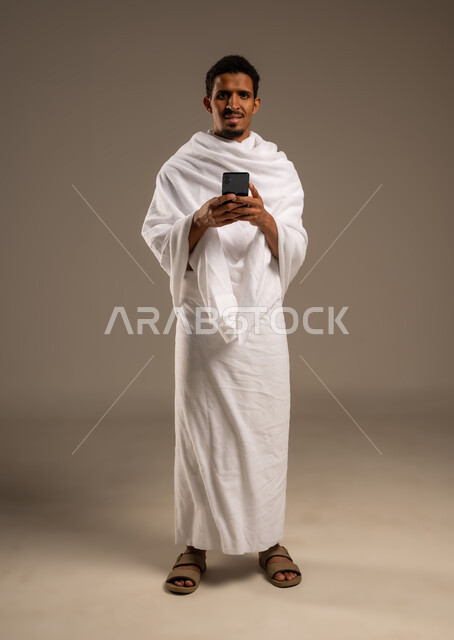 Display of Hajj and Umrah guide applications and programs, portrait of a young Gulf Arab Omani man wearing Ihram clothes holding a mobile phone, use of modern technical devices in communication, full body photo, beige background