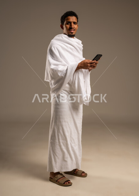 Display of Hajj and Umrah guide applications and programs, portrait of a young Gulf Arab Omani man wearing Ihram clothes holding a mobile phone, use of modern technical devices in communication, full body photo, beige background