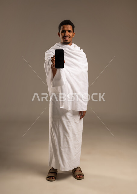 Display of Hajj and Umrah guide applications and programs, portrait of a young Gulf Arab Omani man wearing Ihram clothes holding a mobile phone, use of modern technical devices in communication, full body photo, beige background