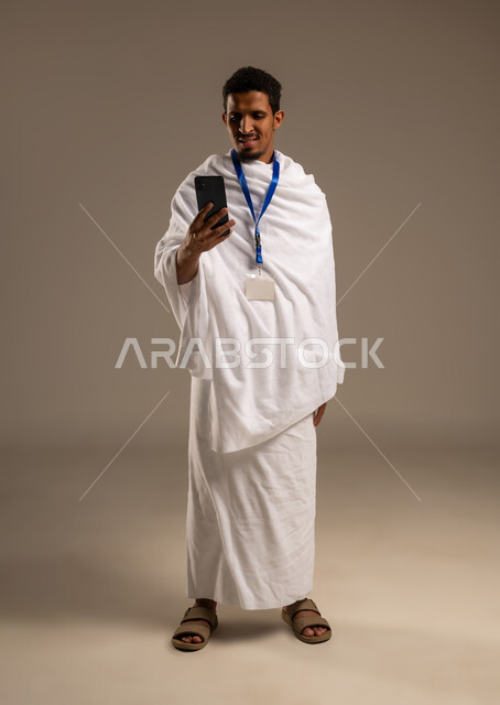 Display of Hajj and Umrah guide applications and programs, portrait of a young Gulf Arab Omani man wearing Ihram clothes holding a mobile phone, use of modern technical devices in communication, full body photo, beige background