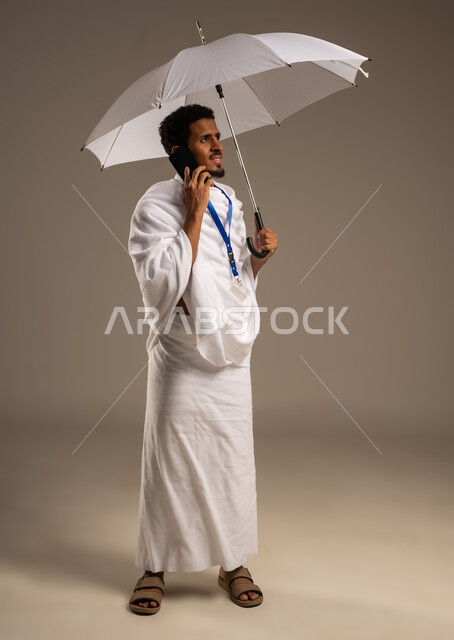 Communicating with family and friends using a mobile phone, portrait of a young Gulf Arab Omani man wearing Ihram clothes making a call using his mobile device holding an umbrella, full body photo, beige background