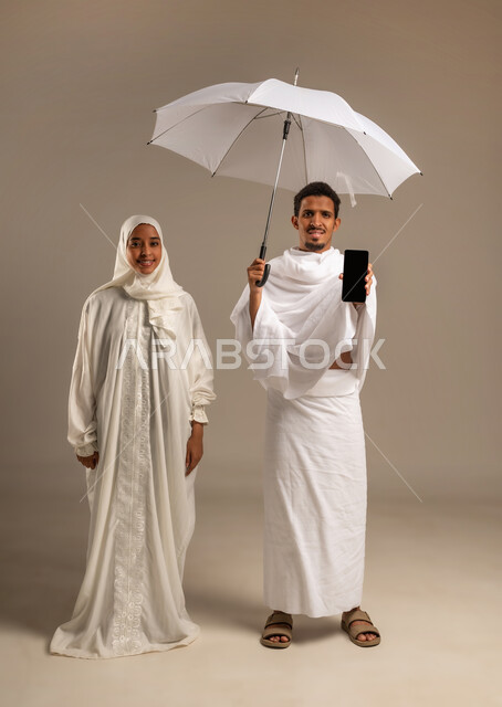 استخدام وسائل التواصل الاجتماعي، الاستعداد لاداء مناسك الحج والعمره, بورتريه لزوجين عربيين خليجيين عمانيين يرتديان ثياب الاحرام يشيران بشاشه الهاتف المحمول الى الامام، التقرب الى الله تعالى، صورة الجسم بالكامل, خلفية بيج