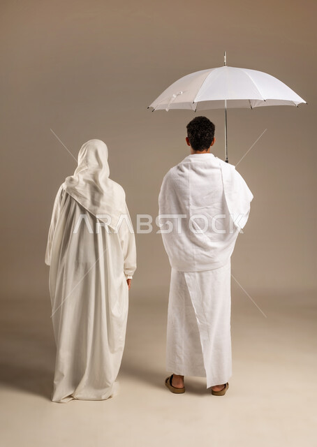 Using a sun umbrella for protection and shade, portrait of an Arab Gulf Omani couple wearing ihram clothes standing straight holding a sun umbrella, getting closer to God Almighty, performing Umrah in the holy month of Ramadan, full body image from the back, beige background
