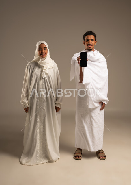 استخدام وسائل التواصل الاجتماعي، الاستعداد لاداء مناسك الحج والعمره, بورتريه لزوجين عربيين خليجيين عمانيين يرتديان ثياب الاحرام يشيران بشاشه الهاتف المحمول الى الامام، التقرب الى الله تعالى، صورة الجسم بالكامل, خلفية بيج