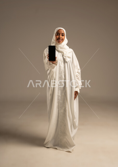 Gestures indicating happiness in performing Hajj and Umrah rituals, using modern technology to communicate with family and friends, portrait of a young Arab Gulf Muslim woman wearing Ihram clothes holding her mobile phone, Hajj season 2025, the fifth pillar of Islam, white background