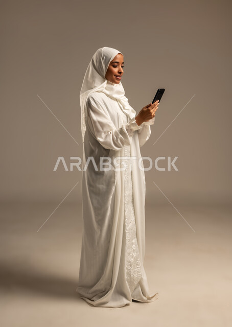 إيماءات تدل على السعادة بأداء مناسك الحج و العمرة, استخدام التقنيات الحديثه للتواصل مع الاهل والاصدقاء, بورتريه لشابة مسلمة خليجية عربية ترتدي ملابس الاحرام تحمل بيدها هاتفهها المحمول ، موسم الحج 2025، الركن الخامس من أركان الاسلام، خلفيه بيضاء