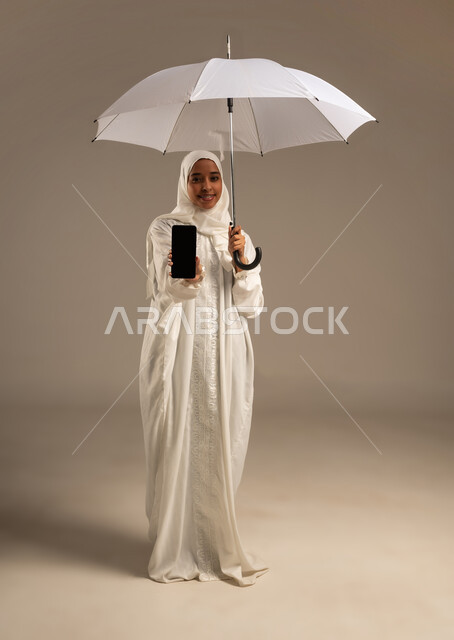 إيماءات تدل على السعادة بأداء مناسك الحج و العمرة, التواصل مع الاهل والاصدقاء باستخدام التقنيات الحديثه, بورتريه لشابة مسلمة خليجية عربية ترتدي ملابس الاحرام تحمل بيدها مظلة وتقوم باستخدام هاتفهها المحمول، موسم الحج 2025، الركن الخامس من أركان الاسلام، خلفيه بيضاء