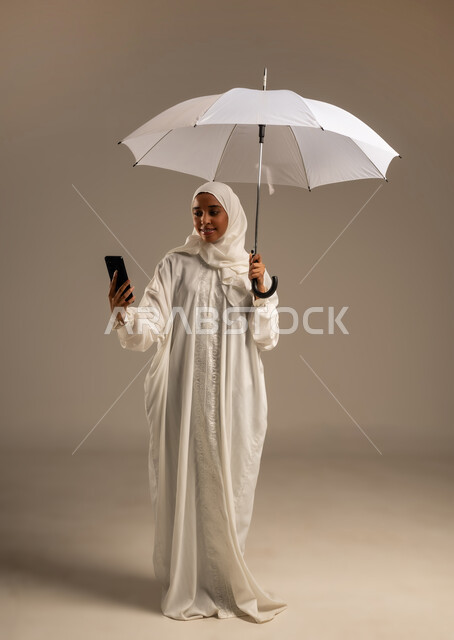 إيماءات تدل على السعادة بأداء مناسك الحج و العمرة, التواصل مع الاهل والاصدقاء باستخدام التقنيات الحديثه, بورتريه لشابة مسلمة خليجية عربية ترتدي ملابس الاحرام تحمل بيدها مظلة وتقوم باستخدام هاتفهها المحمول، موسم الحج 2025، الركن الخامس من أركان الاسلام، خلفيه بيضاء