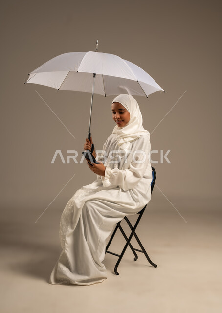 إيماءات تدل على السعادة بأداء مناسك الحج و العمرة, التواصل مع الاهل والاصدقاء باستخدام التقنيات الحديثه, بورتريه لشابة مسلمة خليجية عربية ترتدي ملابس الاحرام تحمل بيدها مظلة وتقوم باستخدام هاتفهها المحمول، موسم الحج 2025، الركن الخامس من أركان الاسلام، خلفيه بيضاء