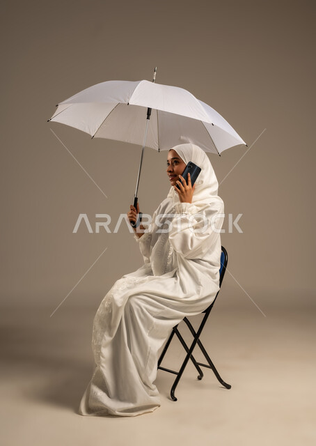 إيماءات تدل على السعادة بأداء مناسك الحج و العمرة, التواصل مع الاهل والاصدقاء باستخدام التقنيات الحديثه, بورتريه لشابة مسلمة خليجية عربية ترتدي ملابس الاحرام تحمل بيدها مظلة وتقوم باستخدام هاتفهها المحمول، موسم الحج 2025، الركن الخامس من أركان الاسلام، خلفيه بيضاء
