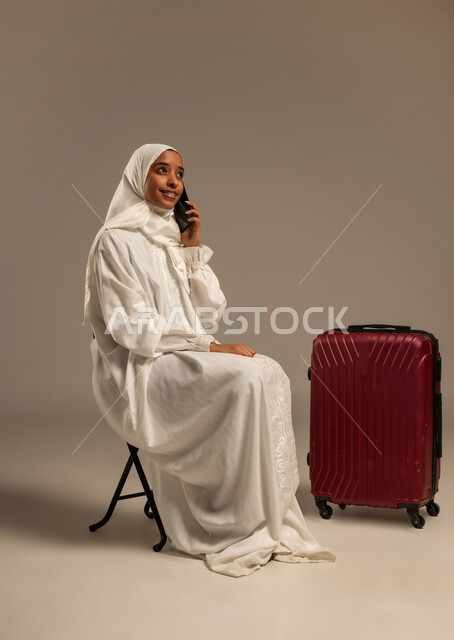 الاستعداد للتوجه و الانطلاق لزيارة بيت الله الحرام، استخدام تطبيقات الجوال للتواصل مع الاهل والاصدقاء, بورتريه لامرأه خليجيه عربيه ترتدي ملابس الاحرام تمسك بيدها هاتفهها المحمول بجوارها حقيبه سفرها, خلفية بيضاء