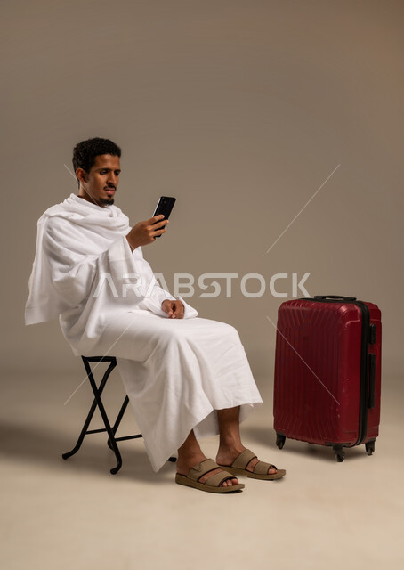 الاستعداد للتوجه و الانطلاق لزيارة بيت الله الحرام، استخدام تطبيقات الجوال للتواصل مع الاهل والاصدقاء, بورتريه لرجل خليجي عربي يرتدي ملابس الاحرام يمسك بيده هاتفهه المحمول بجواره حقيبه سفره, خلفية بيضاء