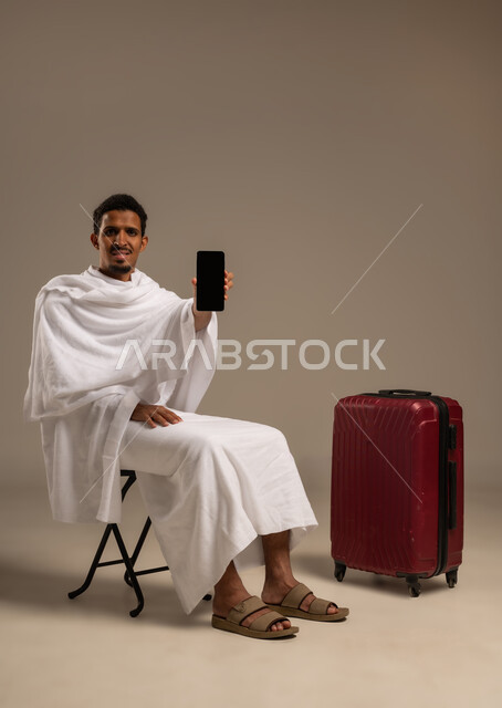 الاستعداد للتوجه و الانطلاق لزيارة بيت الله الحرام، استخدام تطبيقات الجوال للتواصل مع الاهل والاصدقاء, بورتريه لرجل خليجي عربي يرتدي ملابس الاحرام يمسك بيده هاتفهه المحمول بجواره حقيبه سفره, خلفية بيضاء