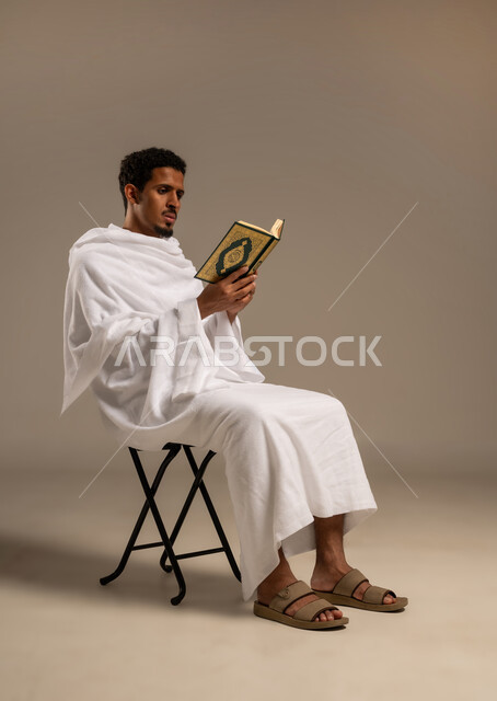 مفهوم العبادة والتقرب إلى الله تعالى، حفظ وتلاوة القرآن الكريم بتدبر وسكينه ، بورتريه  لرجل عربي خليجي يجلس على كرسي يرتدي لباس الإحرام يحمل بيده المصحف الشريف، خلفية بيضاء