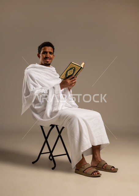 مفهوم العبادة والتقرب إلى الله تعالى، حفظ وتلاوة القرآن الكريم بتدبر وسكينه ، بورتريه  لرجل عربي خليجي يجلس على كرسي يرتدي لباس الإحرام يحمل بيده المصحف الشريف، خلفية بيضاء