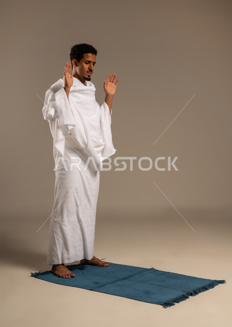 الخشوع في الصلاة والتضرع إلى الله، اداء الركن الثاني من اركان الاسلام الخمسه, بورتريه لرجل عربي خليجي يرتدي ملابس الإحرام يؤدي فريضة الصلاة على سجادته الخاصه, مناسك الحج والعمرة، التقرب إلى الله بالطاعه والعبادات، خلفية بيضاء
