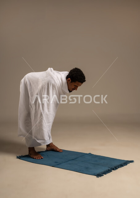 الخشوع في الصلاة والتضرع إلى الله، اداء الركن الثاني من اركان الاسلام الخمسه, بورتريه لرجل عربي خليجي يرتدي ملابس الإحرام يؤدي فريضة الصلاة على سجادته الخاصه, مناسك الحج والعمرة، التقرب إلى الله بالطاعه والعبادات، خلفية بيضاء