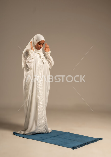 العبادة والتقرب إلى الله، أداء الركن الثاني من أركان الإسلام الخمسة، بورتريه لشابة عربية خليجية ترتدي الحجاب والعبائه البيضاء تؤدي فريضه الصلاه على سجادتها الخاصه بخشوع وسكينه، خلفية بيضاء