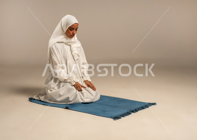 العبادة والتقرب إلى الله، أداء الركن الثاني من أركان الإسلام الخمسة، بورتريه لشابة عربية خليجية ترتدي الحجاب والعبائه البيضاء تؤدي فريضه الصلاه على سجادتها الخاصه بخشوع وسكينه، خلفية بيضاء