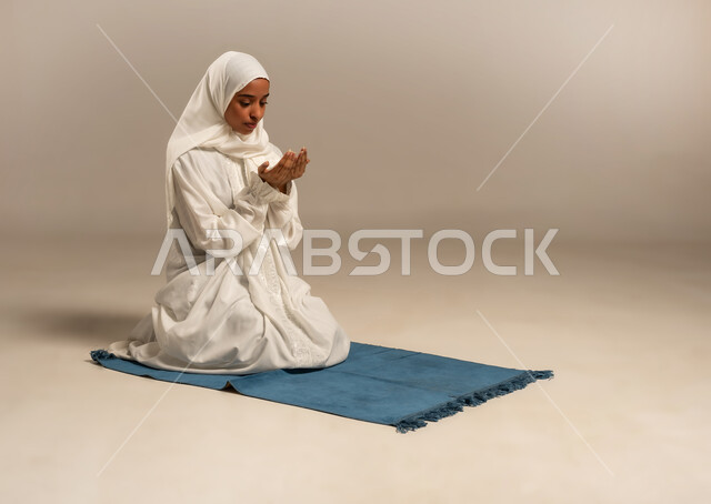 تعابير تدل على التوسل والتضرع، أداء الفروض على وقتها, التقرب الى الله من خلال الإجتهاد في العبادة والحفاظ على الصلوات، بورتريه لإمرأة عربية خليجية ترتدي الحجاب والعباءة البيضاء تجلس على سجادة الصلاة ترفع يديها وتدعو الله، خلفية بيضاء
