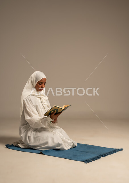 التقرب الى رب العالمين في شهر رمضان المبارك، تلاوة وقراءة كتاب الله,الإجتهاد و الخشوع في العباده, بورتريه لامرأة عربية خليجية محجبة ترتدي العباءة البيضاء تجلس على سجادة الصلاة تمسك بيدها القرآن الكريم, خلفية بيضاء