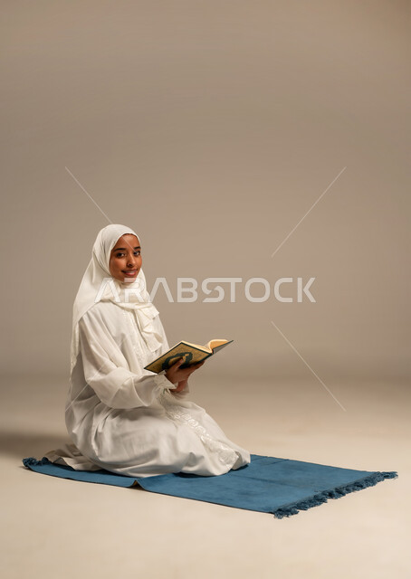 Getting closer to the Lord of the Worlds in the blessed month of Ramadan, reciting and reading the Book of God, diligence and humility in worship, portrait of a veiled Arab Gulf woman wearing a white abaya sitting on a prayer rug holding the Holy Quran in her hand, white background