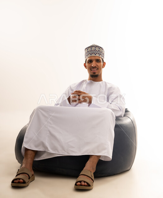 مفهوم الاناقة والاهتمام بالمظهر الخارجي, الاسترخاء والاستمتاع بالأجواء الهادئة، بورتريه لرجل عربي خليجي عماني يرتدي الدشداشه والكمه يجلس على كرسي مريح ينظر الى الكاميرا بايماءات السعادة والسرور، خلفية بيضاء