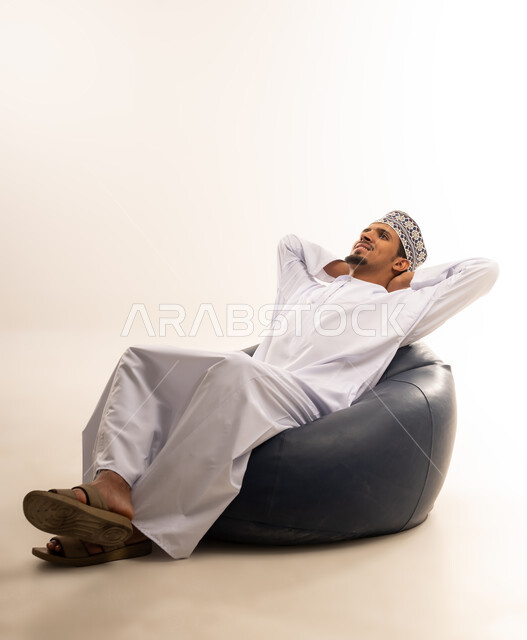 الاسترخاء والاستمتاع بالأجواء الهادئة، مفهوم الاناقة والاهتمام بالمظهر الخارجي, بورتريه لرجل عربي خليجي عماني يرتدي الدشداشه والكمه يجلس على كرسي مريح ينظر الى الاعلى بايماءات الراحه والخلود الى النوم، خلفية بيضاء