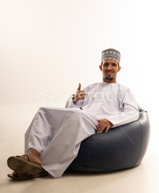 الشعور بالرضا والقبول، مفهوم النجاح وتحقيق الأهداف، بورتريه لشاب عربي خليجي عماني يرتدي الدشداشة والكمه يجلس على كرسي مريح و ينظر الى الكاميرا رافعا اصبع الابهام للاعلى بايماءات الجودة والامتياز، تعابير الفرح والسرور، خلفية بيضاء