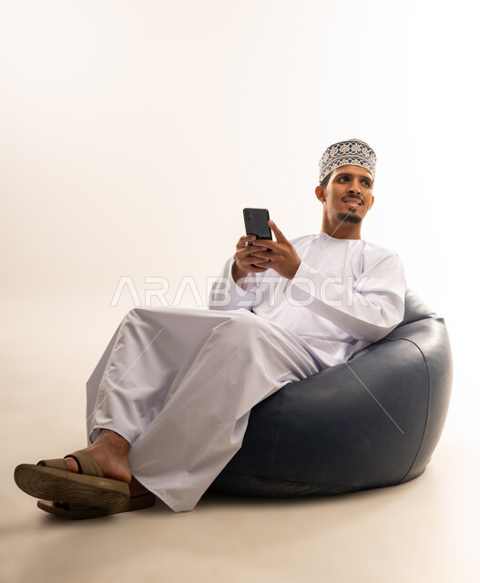 الدردشة مع الاهل والأصدقاء عبر تطبيقات الجوال، تصفح مواقع التواصل الاجتماعي, بورتريه لرجل عربي خليجي عماني يرتدي الدشداشة والكمه يجلس على كرسي مريح يحمل بيده الهاتف المحمول ينظر الى مكان ما, استخدام التقنيات الحديثة المتطورة, خلفية بيضاء